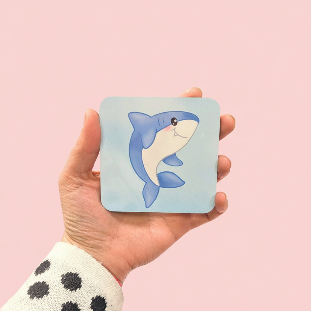 A hand holding a square coaster with a shark design on it.