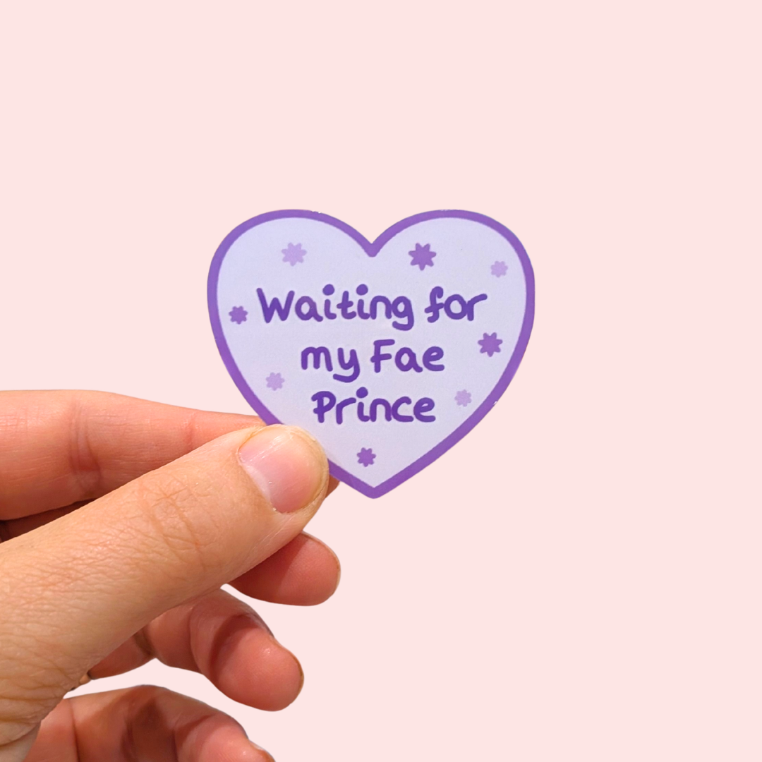 A hand holding a heart-shaped sticker with a light purple background, dark purple border and text that reads 'Waiting for my Fae Prince' with small star accents, against a solid pale pink background.