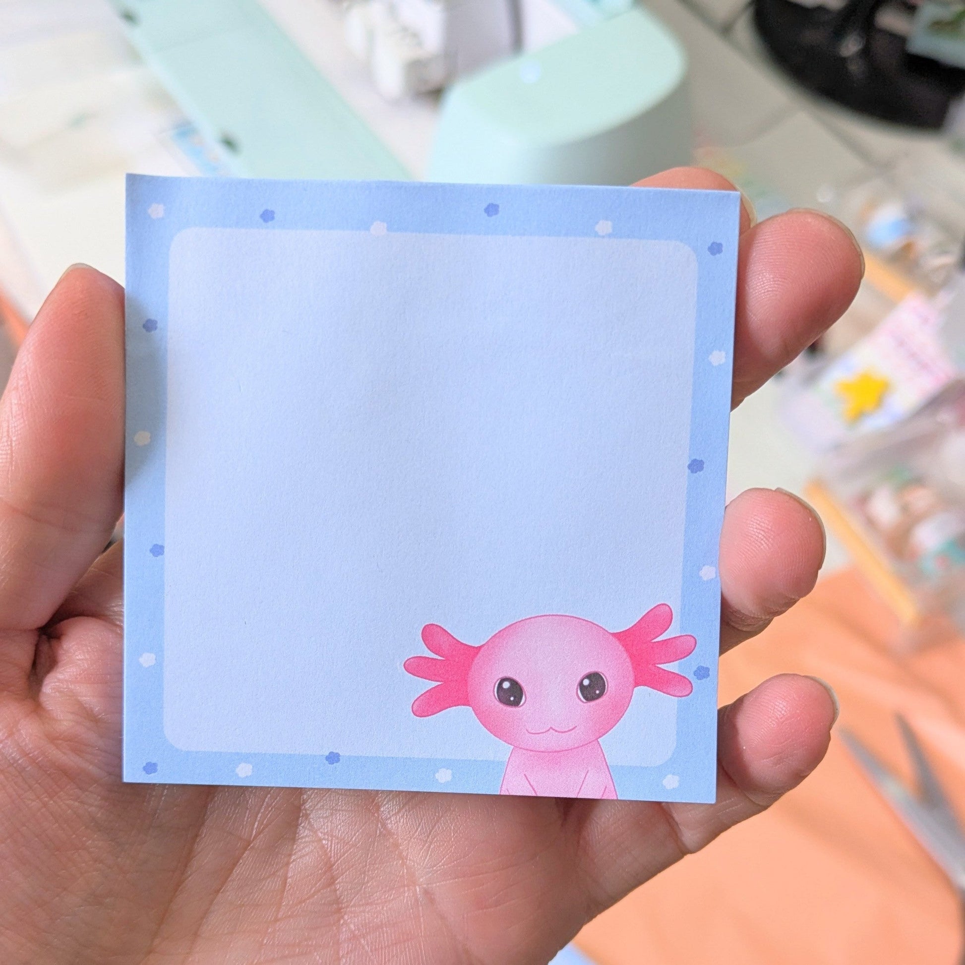 A hand holding a square blue sticky note with an illustration of Axel the Axolotl on it 