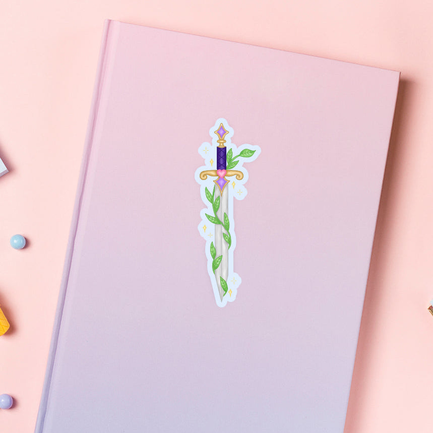 Notebook with a fantasy style sword wrapped in vines sticker on a pink background