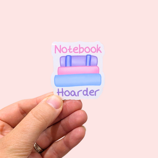 A hand holding a die-cut sticker that reads 'Notebook Hoarder' with pastel-colored illustrated notebooks on a light pink background.