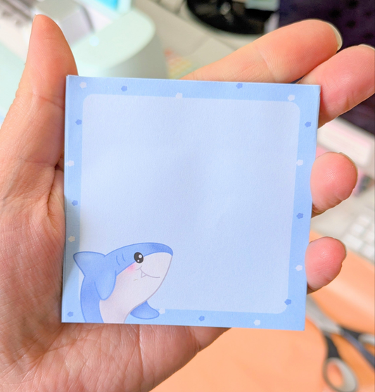 A hand holding a blue sticky note with a cute shark illustration on it.
