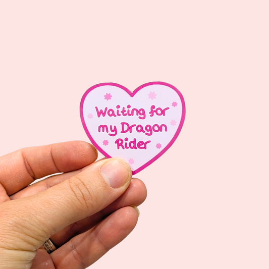 A hand holding a heart-shaped sticker with the text 'Waiting for my Dragon Rider' in pink lettering on a light pink background with a darker pink heart border.